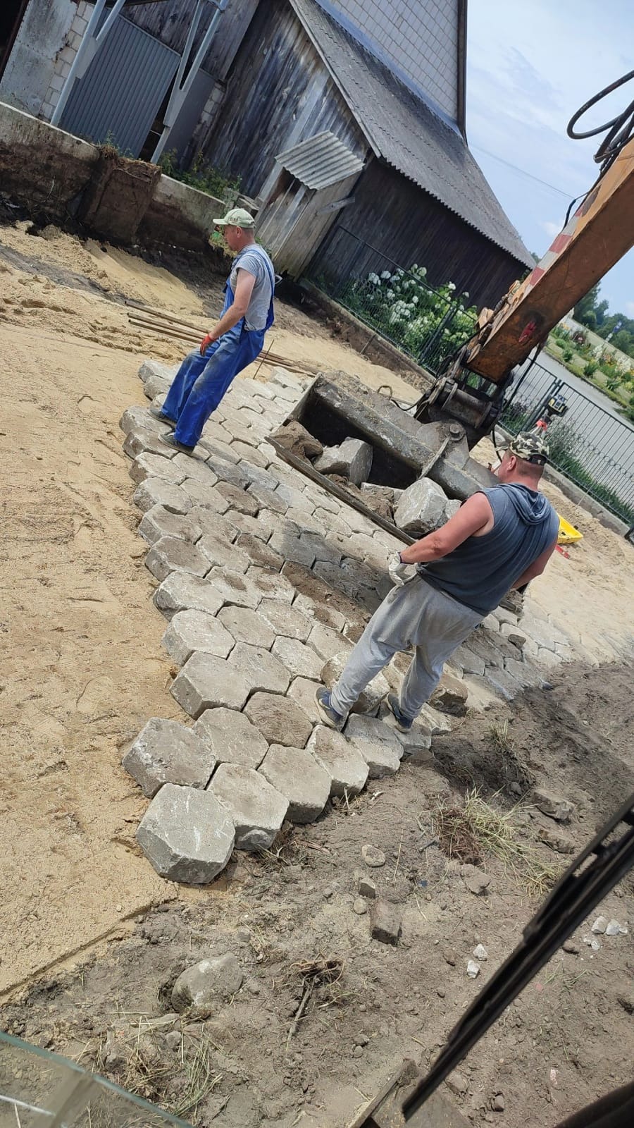 Dwóch pracowników układa szare, sześciokątne płyty chodnikowe na piaszczystym podłożu, przy użyciu koparki w tle, widoczny fragment budynku gospodarczego.