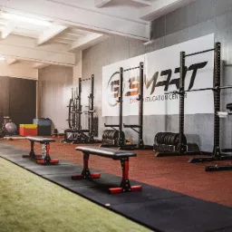 Widok wnętrza siłowni z różnorodnym sprzętem do ćwiczeń siłowych, w tle logo 'SMART Sports Education Center' na ścianie, gumowa podłoga i sztuczna trawa.