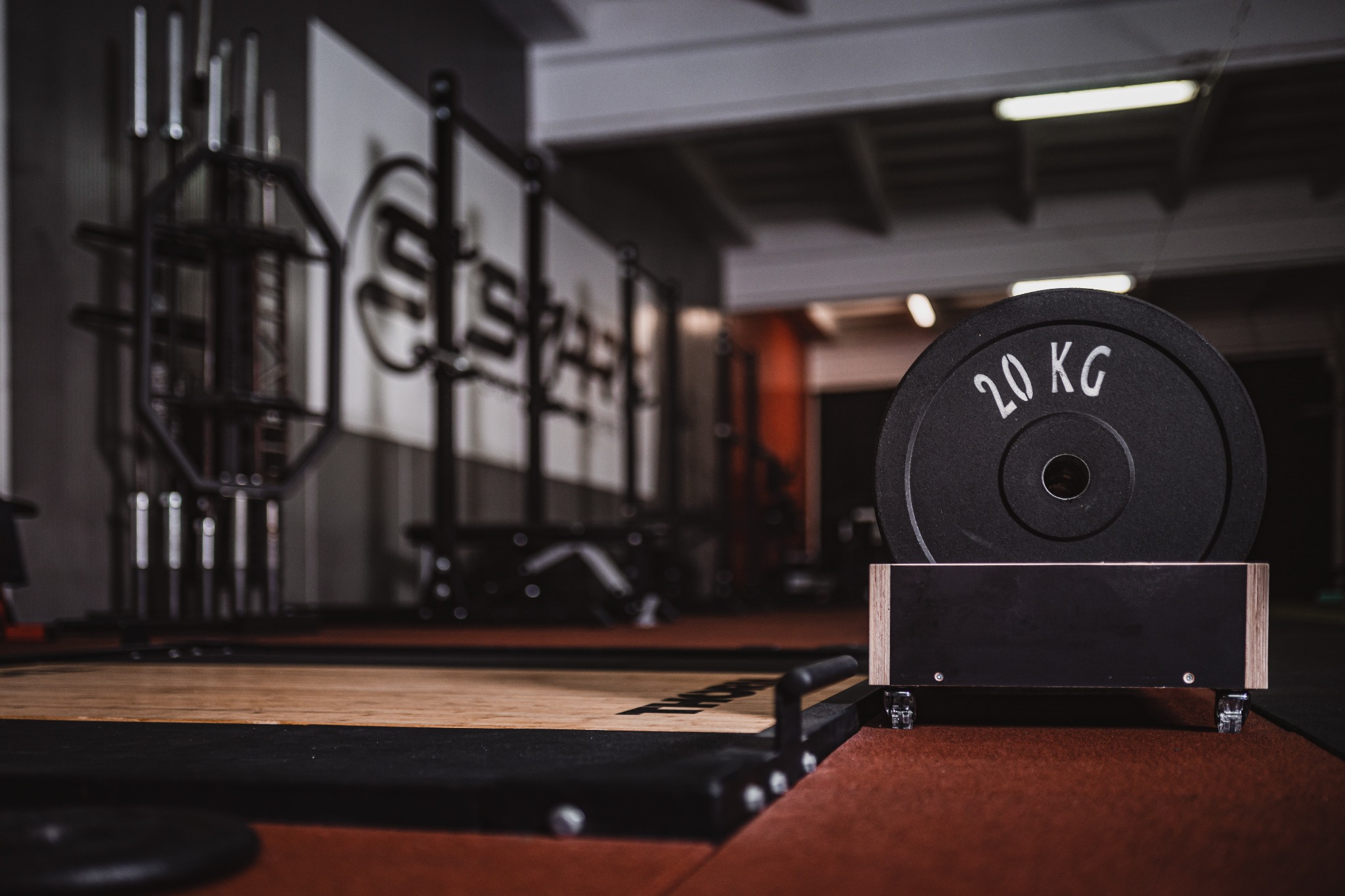 Sala treningowa z platformą do podnoszenia ciężarów i stojakiem na obciążenia z jednym czarnym talerzem 20 kg na pierwszym planie, w tle widoczne klatki do ćwiczeń i logo firmy.