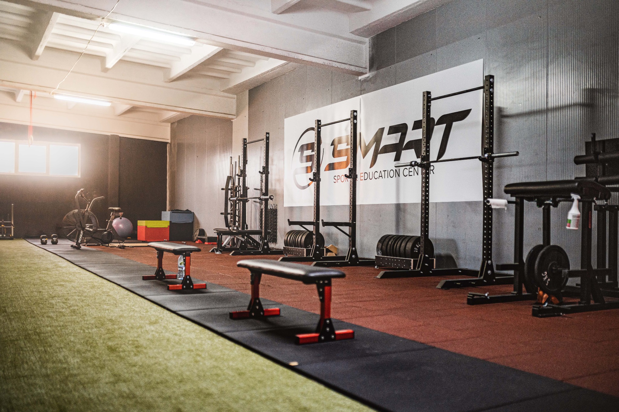 Widok wnętrza siłowni z różnorodnym sprzętem do ćwiczeń siłowych, w tle logo 'SMART Sports Education Center' na ścianie, gumowa podłoga i sztuczna trawa.