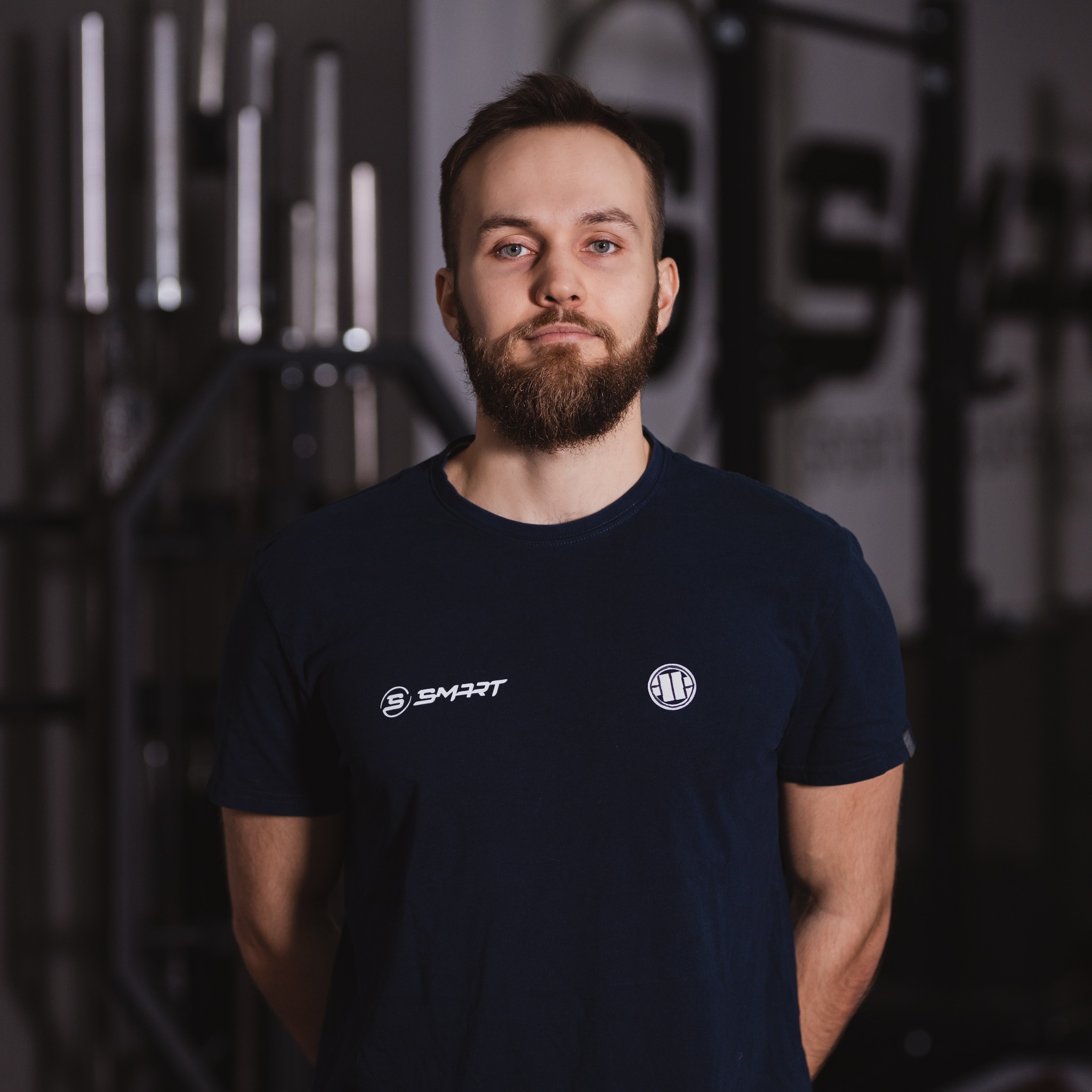 Portret brodatego mężczyzny w granatowej koszulce z logo na tle siłowni z hantlami i sztangami.