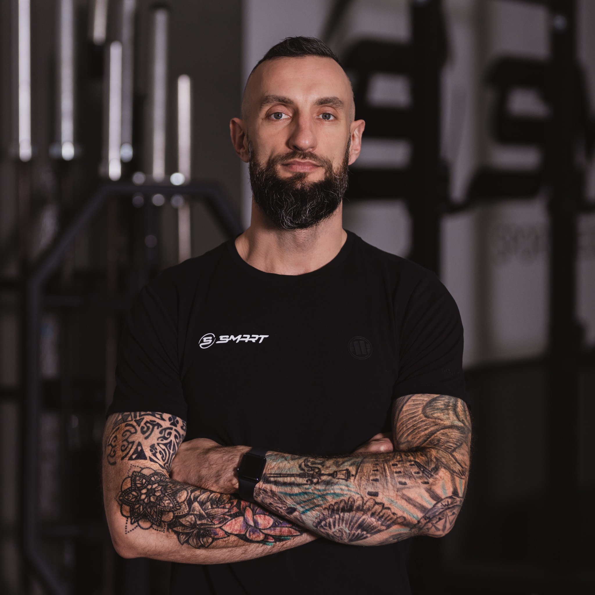 Mężczyzna z brodą i tatuażami na ramionach, ubrany w czarną koszulkę z logo, stoi na tle sprzętu do ćwiczeń.