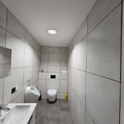 Wnętrze łazienki wykończonej szarą, wielkoformatową płytką z fugą w kolorze czarnym, widok na umywalkę z szafką, pisuar, sedes podwieszany i oświetlenie sufitowe LED.