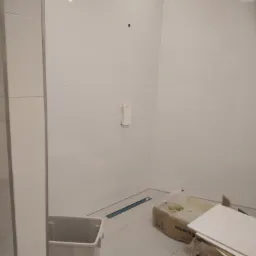 Łódź: Wnętrze łazienki w trakcie wykańczania, widoczne białe płytki na ścianach, karton z płytami Rockfon, odpływ liniowy w podłodze i plastikowy kosz na śmieci.