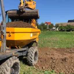 Żółta wywrotka Terex z pełną łyżką ziemi na tle zielonego wzgórza i zabudowań, przygotowująca teren pod budowę.