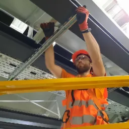 Elektryk w pomarańczowej kamizelce i kasku, montuje koryto kablowe pod sufitem, używając klucza. Widoczne perforowane korytka kablowe i elementy mocujące.