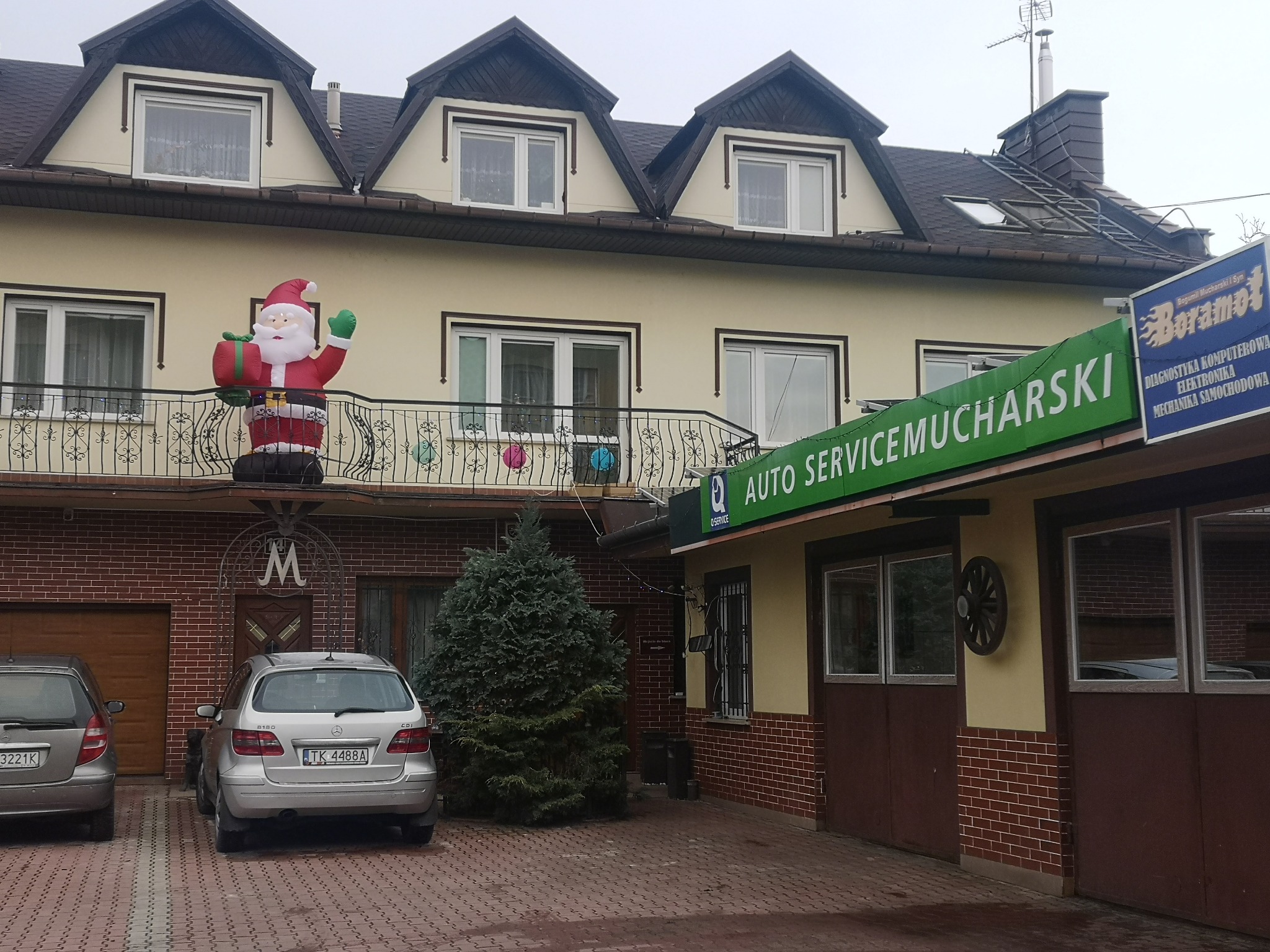 Budynek warsztatu samochodowego w Kielcach z dwoma zaparkowanymi Mercedesami, na balkonie dmuchany Święty Mikołaj, a nad wejściem zielony szyld 'Auto Service Mucharski' i niebieski szyld 'Boramot'...