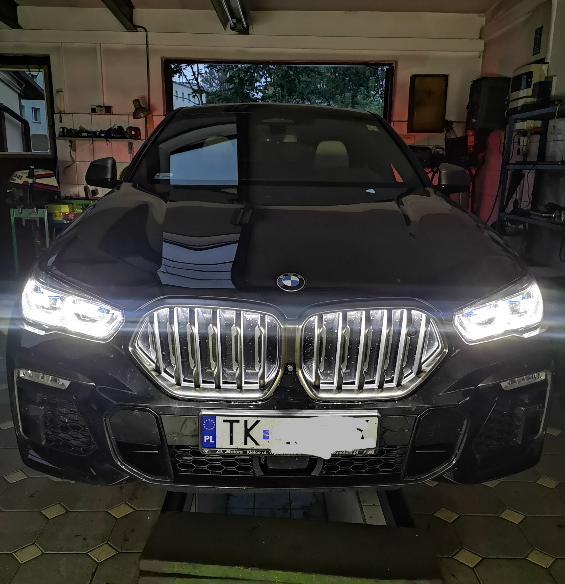 Przód ciemnego BMW X6 z włączonymi światłami, stojącego w warsztacie samochodowym na podnośniku, widoczne narzędzia i wyposażenie warsztatu w tle.