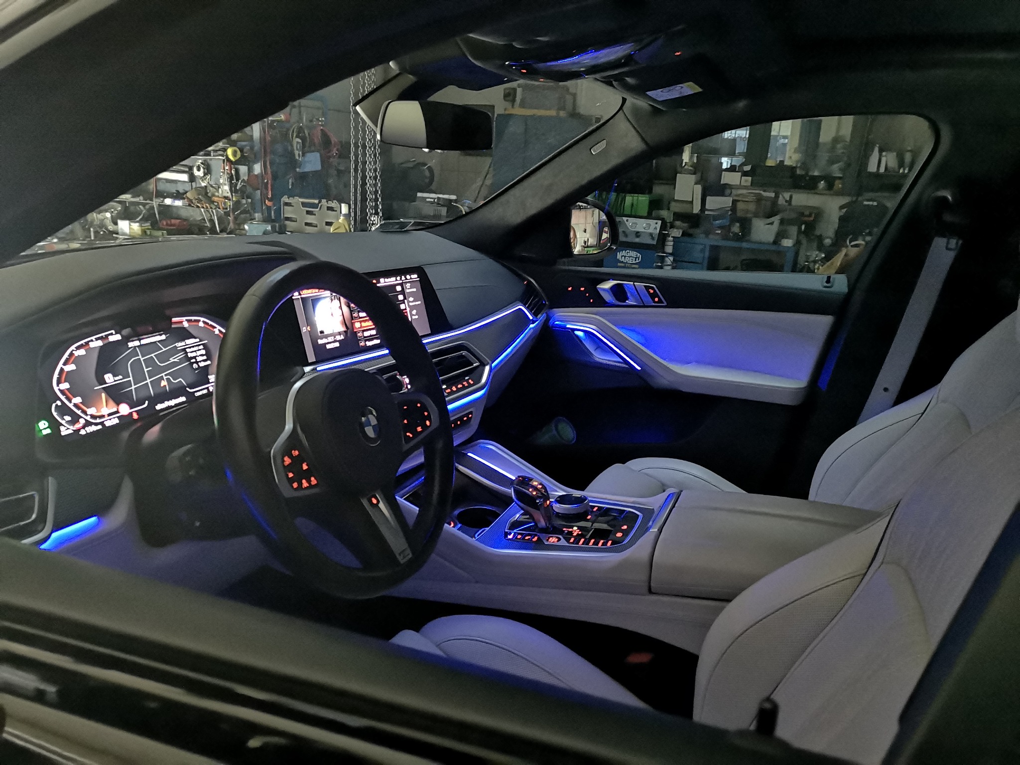 Wnętrze samochodu BMW z białymi skórzanymi siedzeniami i niebieskim podświetleniem ambientowym, widok z zewnątrz przez otwarte drzwi, w tle warsztat samochodowy.