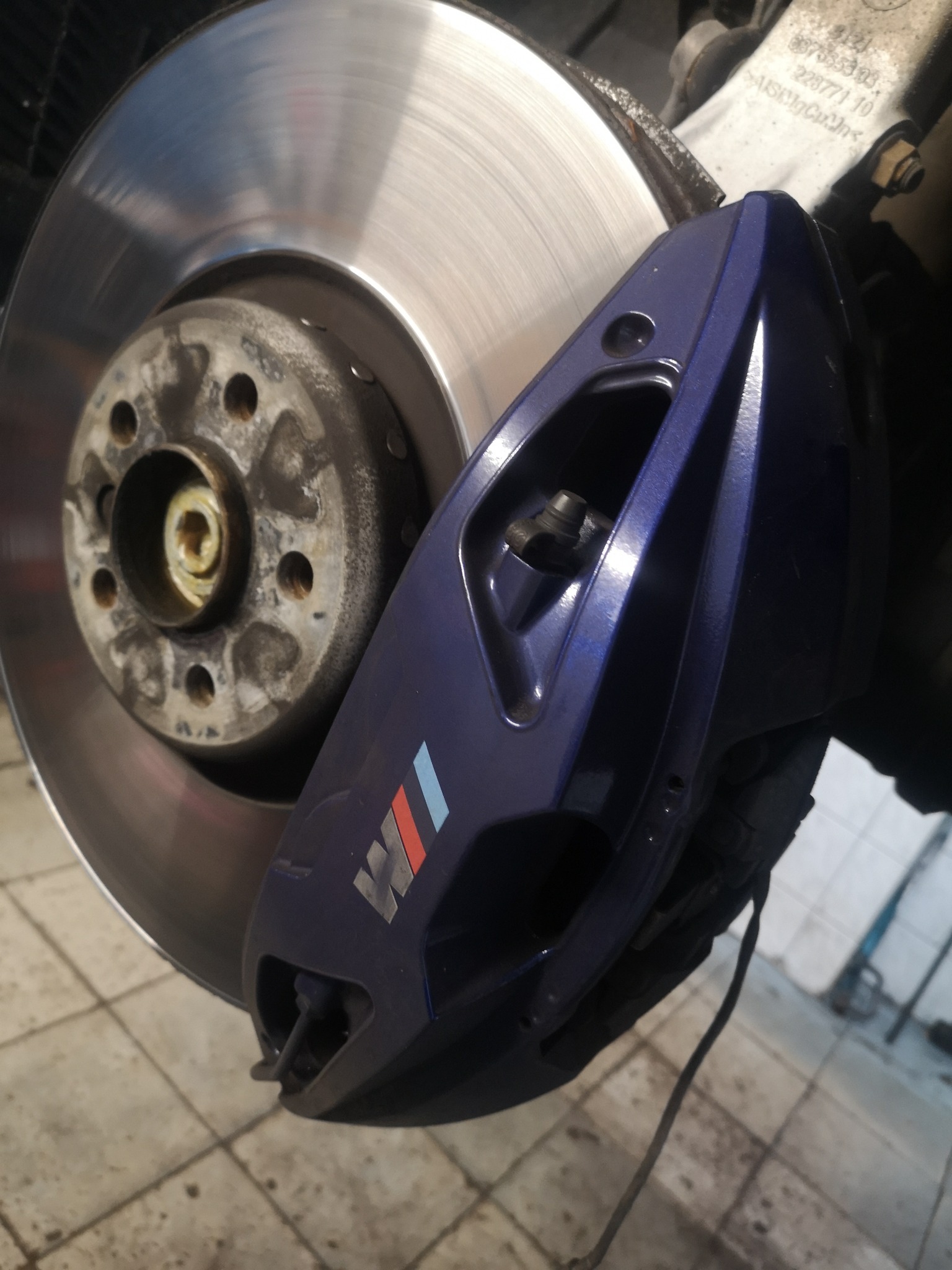 Niebieski zacisk hamulcowy z logo M na tle tarczy hamulcowej, widoczne ślady użytkowania na elementach.