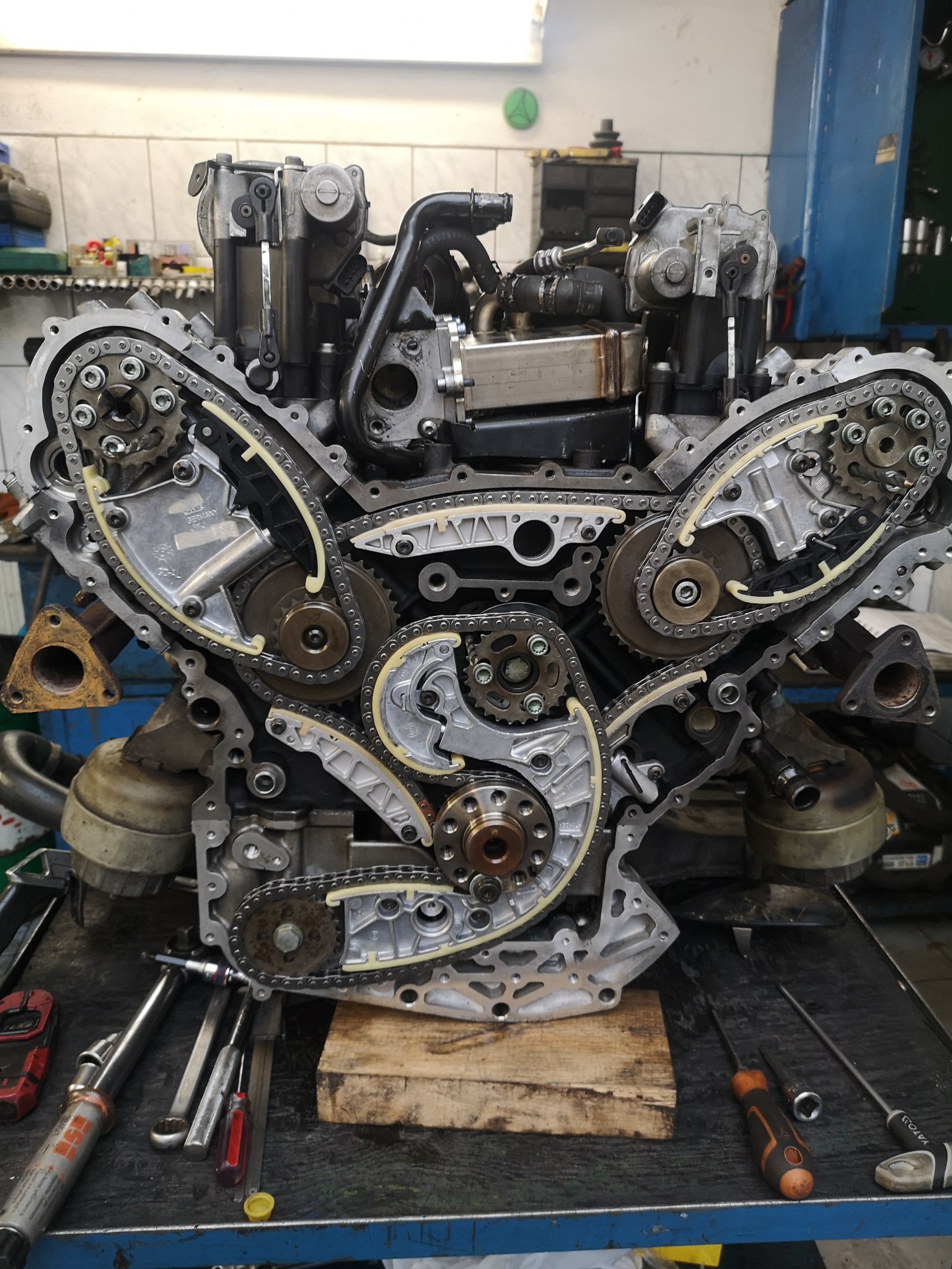 Zdemontowany silnik samochodowy V-kształtny z widocznymi łańcuchami rozrządu i kołami zębatymi, umieszczony na warsztatowym stole roboczym z narzędziami w tle.