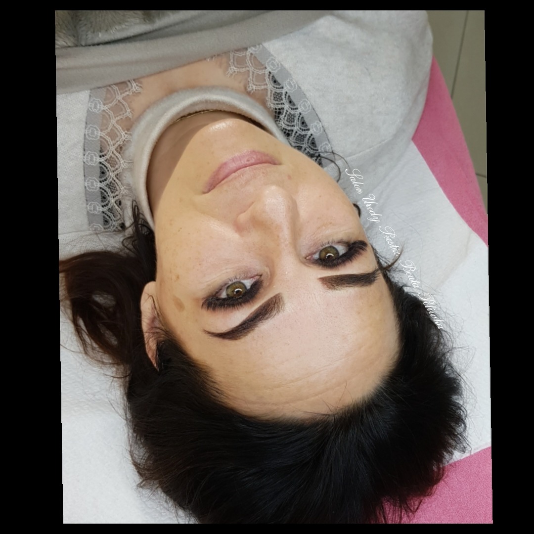 Kobieta z widocznymi, świeżo wykonanymi, ciemnymi brwiami permanentnymi i sztucznymi rzęsami, leżąca na białym podkładzie z różowym akcentem.