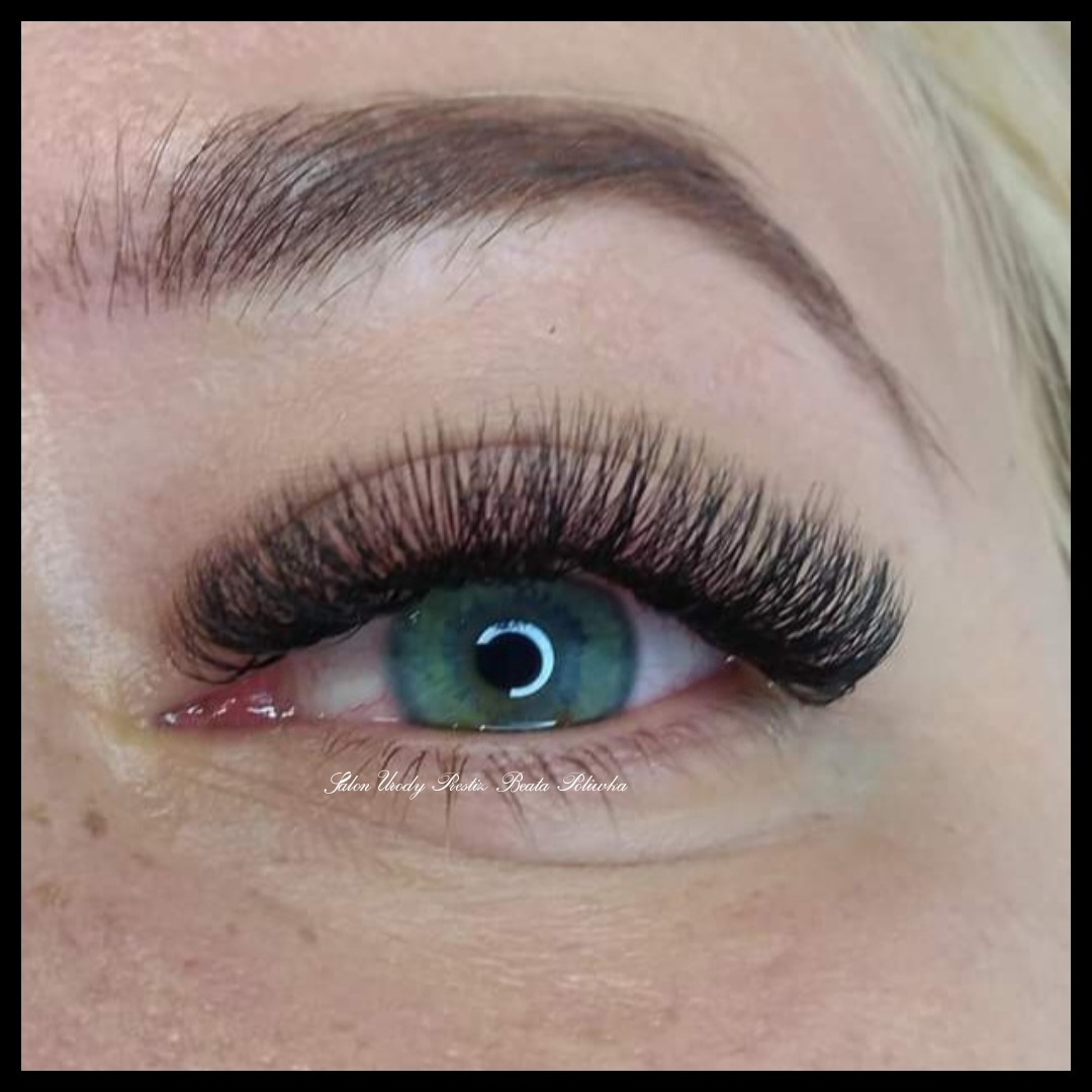 Zbliżenie na oko z zieloną tęczówką i przedłużonymi, gęstymi, czarnymi rzęsami, widoczny delikatny napis 'Salon Urody Rzęs Beata Pilarska' w dolnej części zdjęcia.