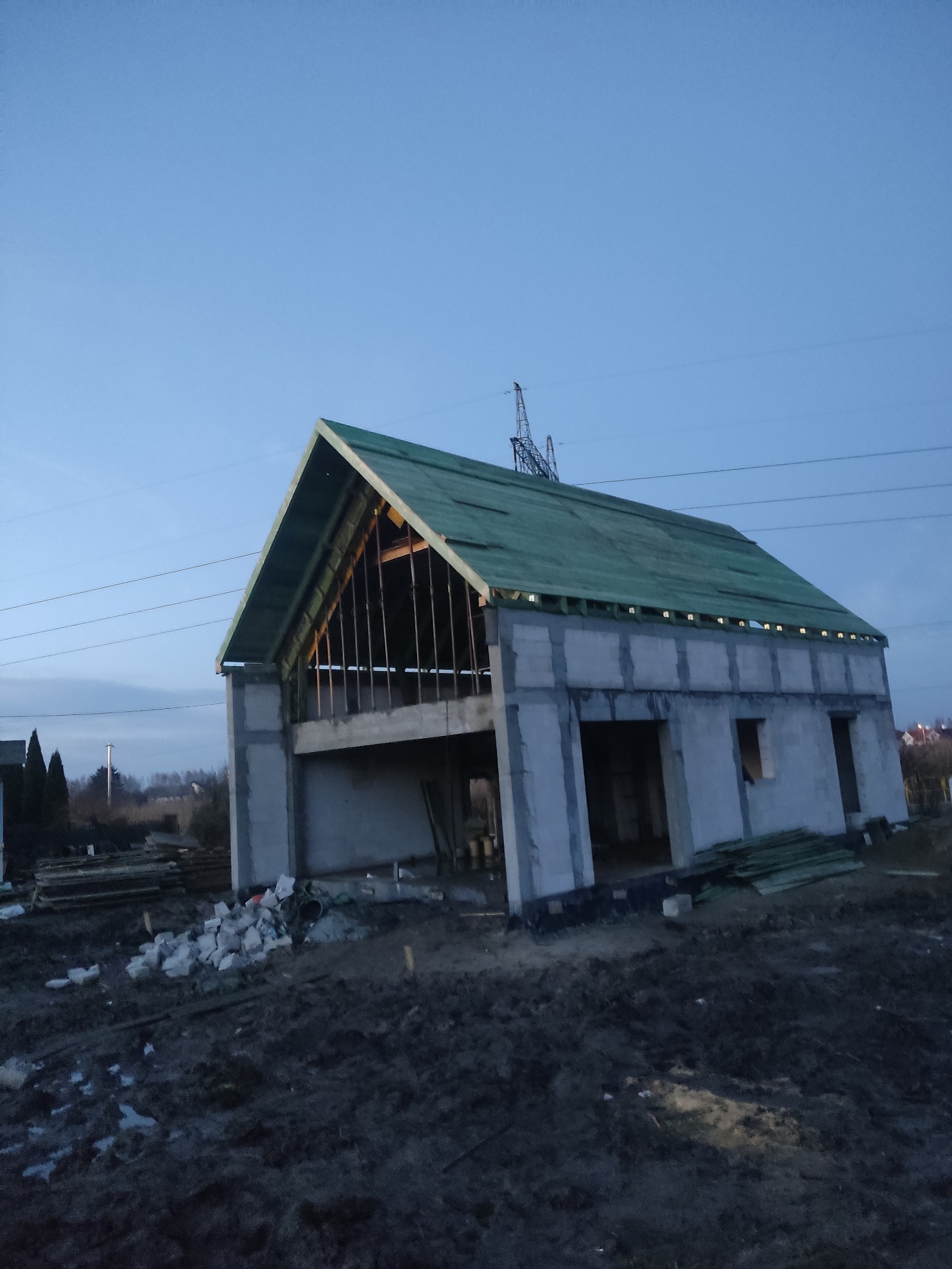 Surowa konstrukcja domu z betonowych bloków i drewnianą konstrukcją dachu pokrytą zieloną membraną dachową, widok z zewnątrz, w tle słupy wysokiego napięcia.