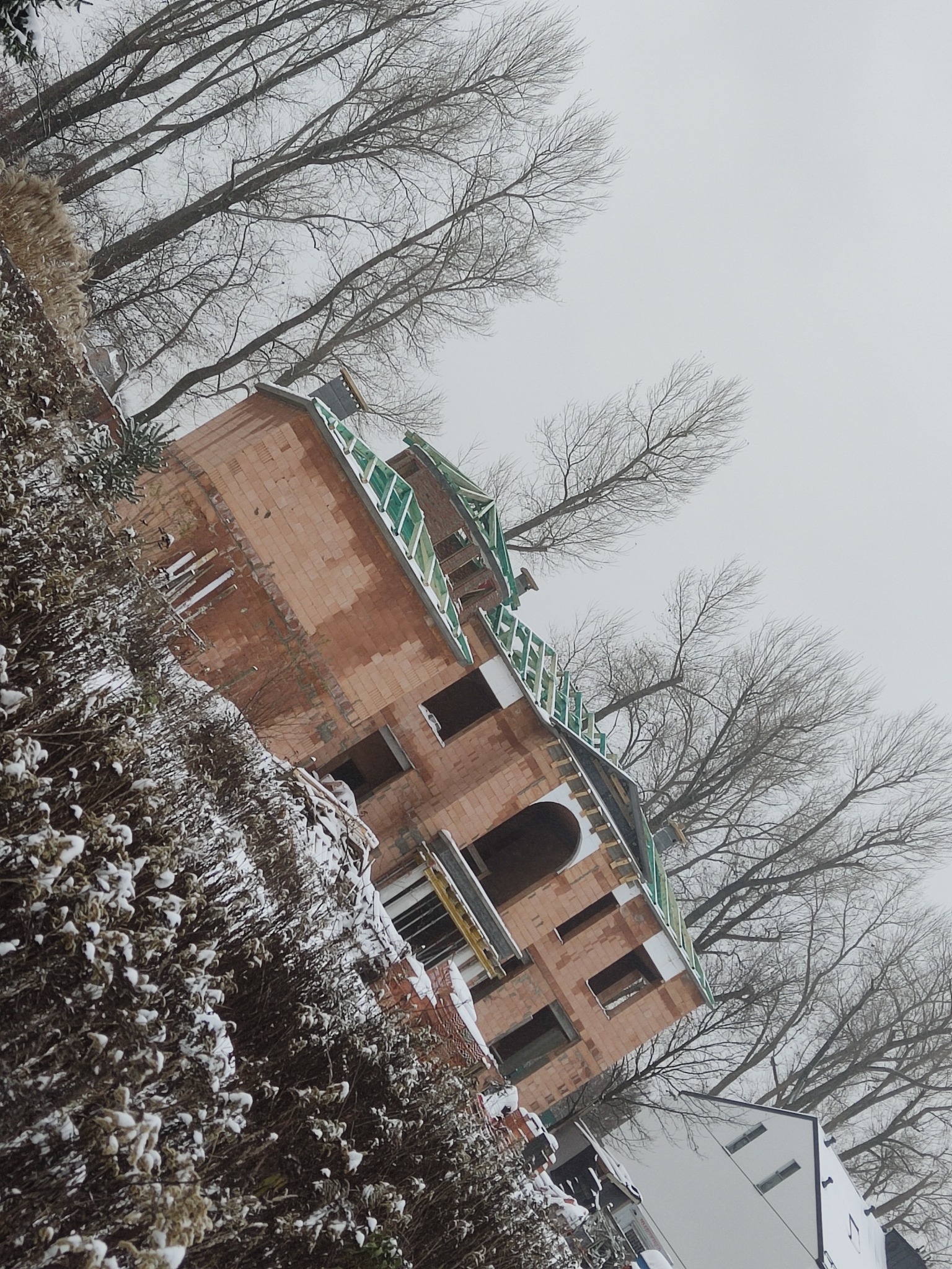 Surowy stan budynku z czerwonej cegły w zimowej scenerii, widoczne elementy konstrukcyjne dachu i okna bez szyb, na pierwszym planie ośnieżone krzewy, w tle nagie drzewa i szare niebo.