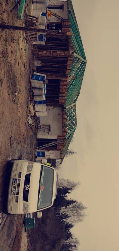 Budowa domu w trakcie montażu konstrukcji dachu, widoczny van firmy na pierwszym planie i materiały budowlane obok