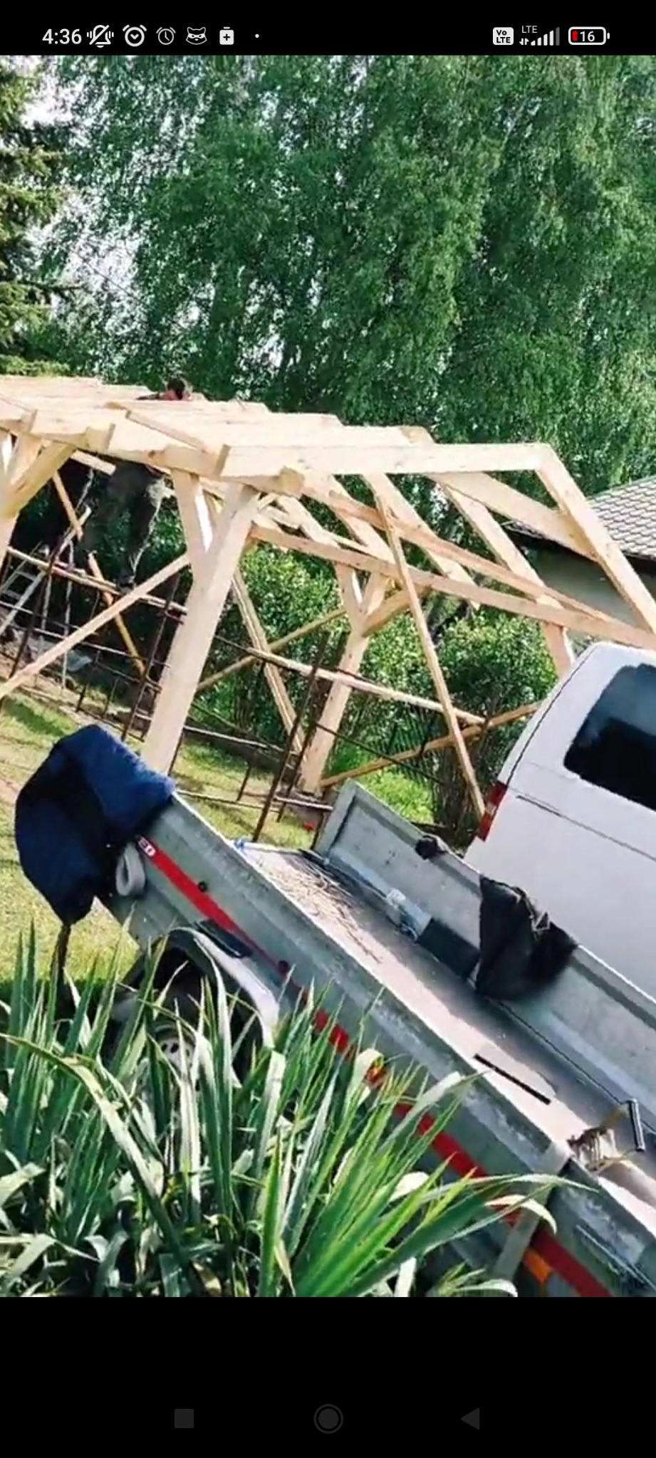 Konstrukcja drewniana z elementami dachu w trakcie montażu, widoczna z perspektywy przyczepy samochodowej, na tle zieleni.