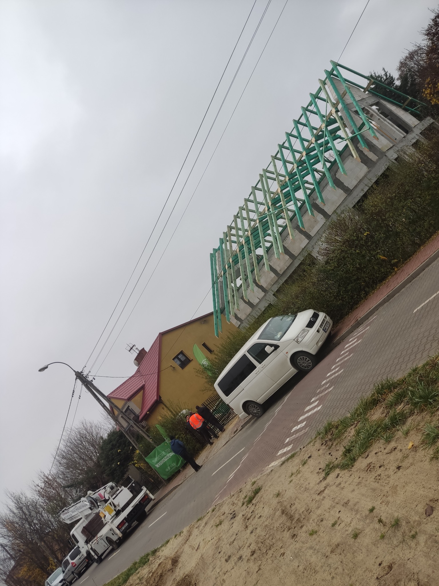 Konstrukcja szkieletowa dachu z impregnowanego drewna, widoczny biały van na tle budowy i żółty dom z czerwonym dachem.