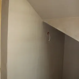 Świeżo nałożony, gładki tynk maszynowy na ścianie wewnętrznej, widoczny fragment wystającego kabla elektrycznego w górnej części ściany i niedoskonałości na podłodze przy ścianie.