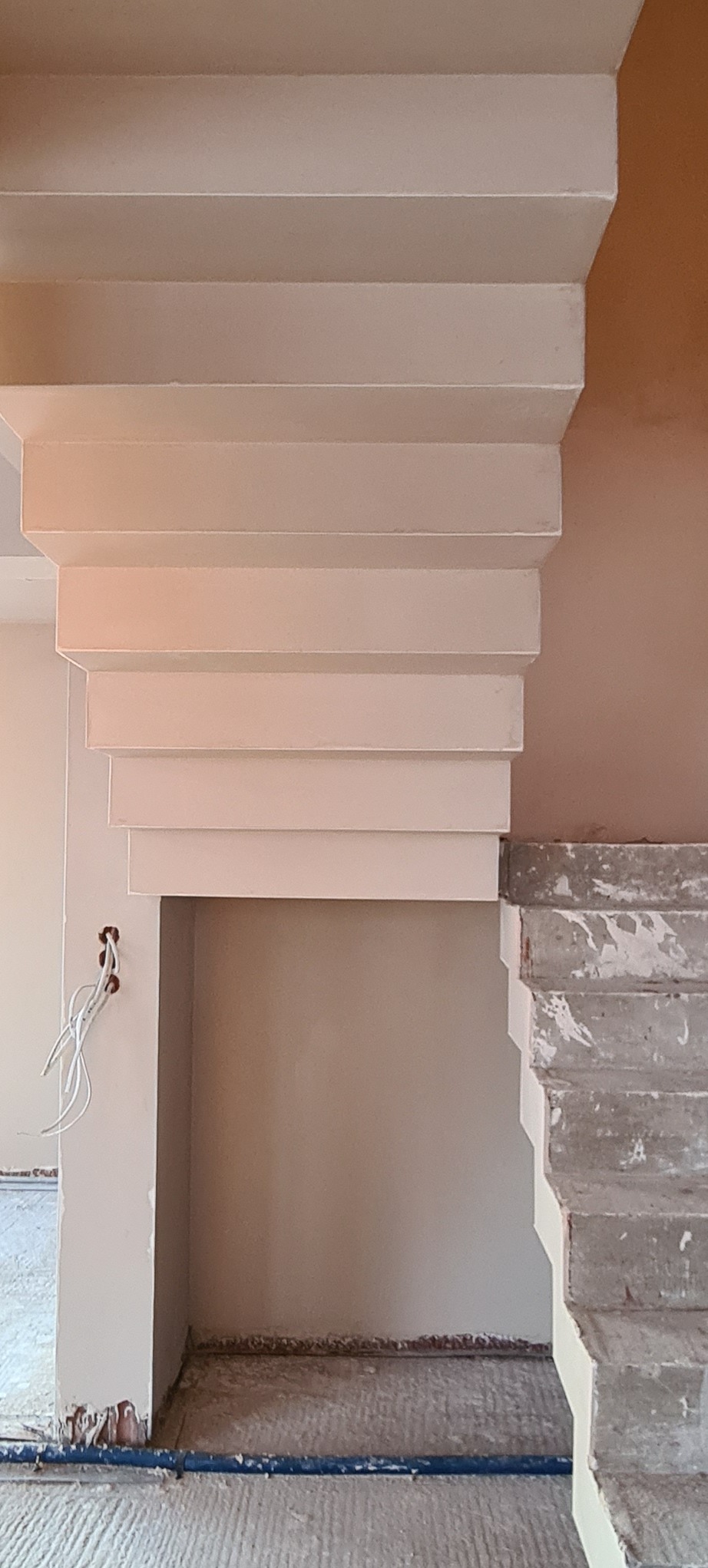 Wykończony fragment sufitu ze schodkowym elementem dekoracyjnym, świeżo po nałożeniu gładzi, z widocznymi przewodami elektrycznymi i surowymi schodami betonowymi w tle.