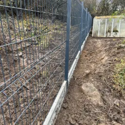 Nowo postawione, szare ogrodzenie panelowe z betonową podmurówką na nierównym, błotnistym terenie. W tle widoczna stara szklarnia i drzewa bez liści.