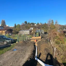 Rów z ułożoną rurą drenażową w białej geowłókninie, w tle traktor z łyżką wypełnioną piaskiem i zabudowania jednorodzinne w słoneczny dzień.