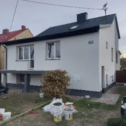 Dwukondygnacyjny dom po tynkowaniu na biało, z ciemnoszarym dachem i numerem 55, widok z boku na ogród, gdzie leżą puste wiadra po farbie.