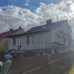 Bas-Bud - Dom w trakcie prac elewacyjnych, widoczne rusztowania, częściowo otynkowana ściana, nowy dach z oknem dachowym, ogrodzenie z siatki i drewnianych sztachet na pierwszym planie.