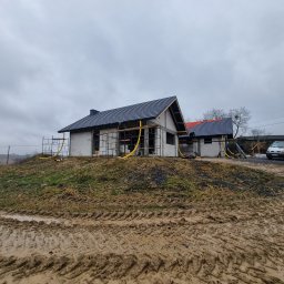 Bas-Bud - Budowa domu jednorodzinnego w trakcie krycia dachu dachówką ceramiczną, widoczne rusztowania i żółte węże wentylacyjne, teren wokół rozjeżdżony przez ciężki sprzęt w pochmurny dzień.
