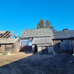 Stary budynek gospodarczy z częściowo zdjętym i częściowo nowym dachem, widoczne etapy prac dekarskich, ziemiste podwórze i błękitne niebo.