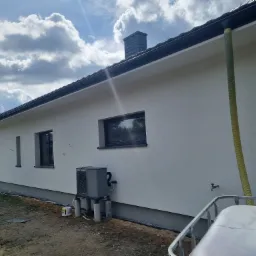 Jasna elewacja domu jednorodzinnego z ciemnymi oknami i szarym kominem, widoczny fragment dachu, rynna i pompa ciepła na zewnątrz budynku, z żółtym wężem przy elewacji.