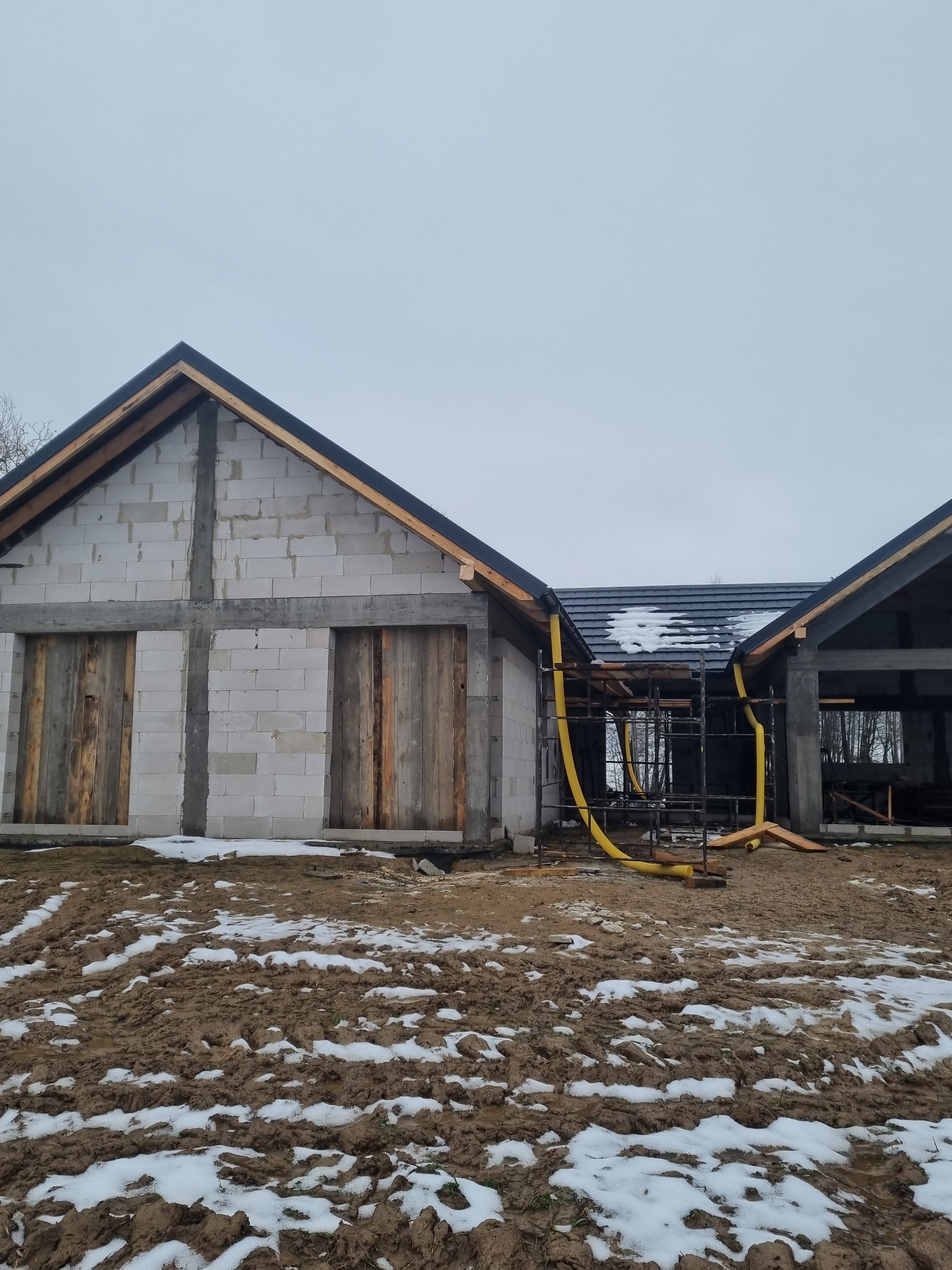 Surowy stan domu z pustaków z częściowo ukończonym dachem z dachówki, żółty przewód wentylacyjny w rusztowaniu, błotniste podłoże z resztkami śniegu.