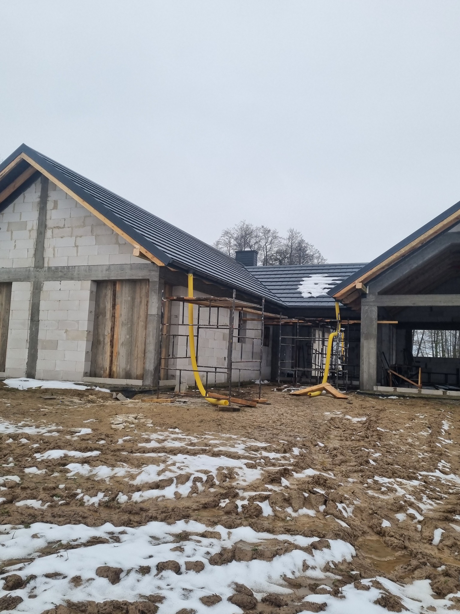 Surowy stan domu jednorodzinnego z czarnym dachem w trakcie budowy, widoczne rusztowania i błotniste podłoże z resztkami śniegu.