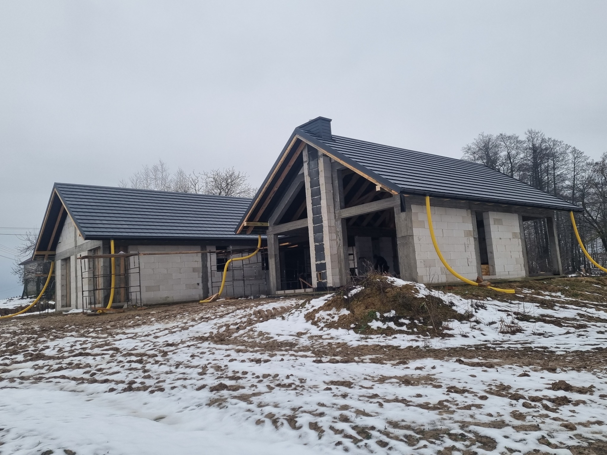 Dwa budynki w stanie surowym zamkniętym z czarną dachówką, widoczne żółte węże wentylacyjne, ziemia pokryta śniegiem, pochmurne niebo.