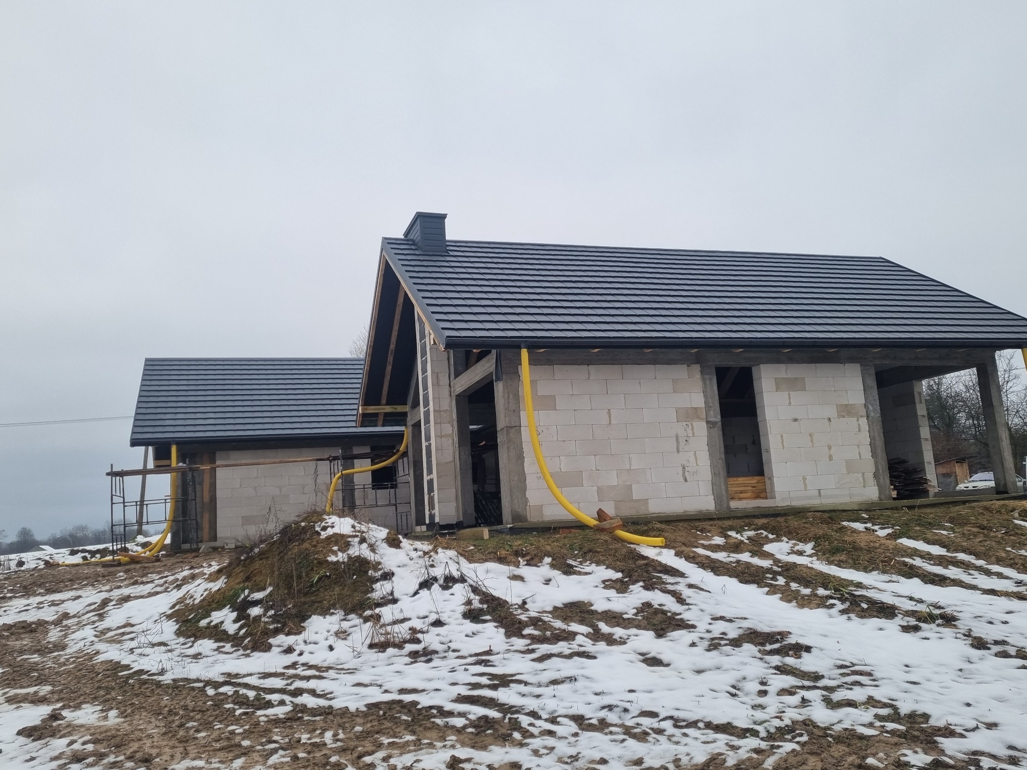 Dwa surowe budynki z szarej cegły z nowymi, ciemnoszarymi dachami z dachówki, widoczne rusztowania i żółte rury na tle zaśnieżonego gruntu.