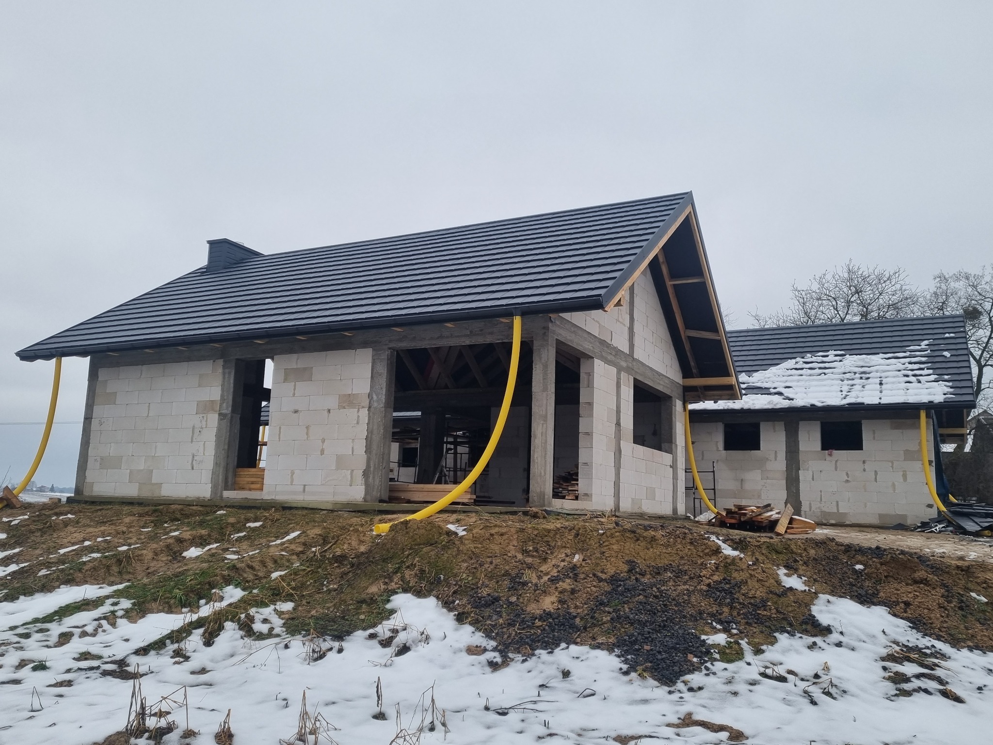 Surowy stan dwóch budynków z czarną dachówką na wzniesieniu pokrytym śniegiem, widoczne żółte rury wentylacyjne