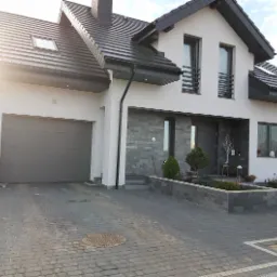 Nowoczesny dom z szarą bramą garażową, elewacją z kamienia dekoracyjnego i kostką brukową na podjeździe w słoneczny dzień.