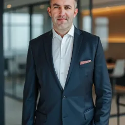 Portret biznesowy mężczyzny w garniturze na tle przeszklonego biura. Widoczna poszetka w kieszonce marynarki i nowoczesne wnętrze biurowe.