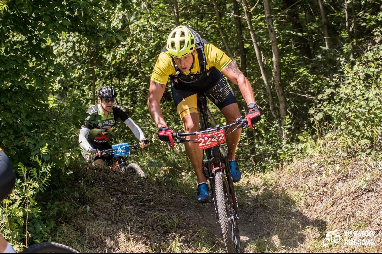 Dwóch mężczyzn na rowerach górskich rywalizuje w zalesionym terenie. Jeden ma żółtą koszulkę, kask i numer 225. Drugi w zielonej koszulce z numerem 42.