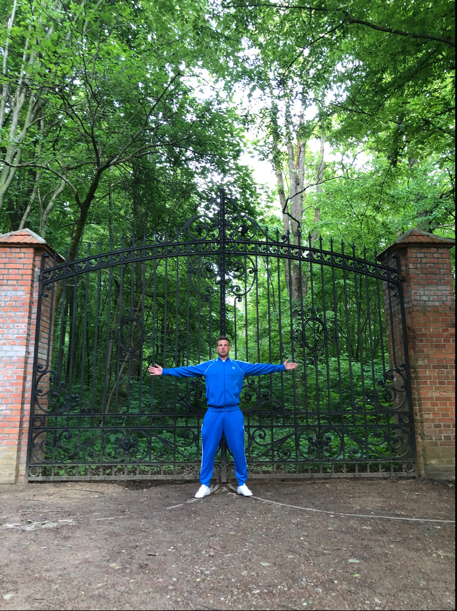 Mężczyzna w niebieskim dresie stoi przed ozdobną, zamkniętą, czarną bramą z ceglanymi słupkami, na tle zielonego lasu. Rozłożone ramiona, białe buty.
