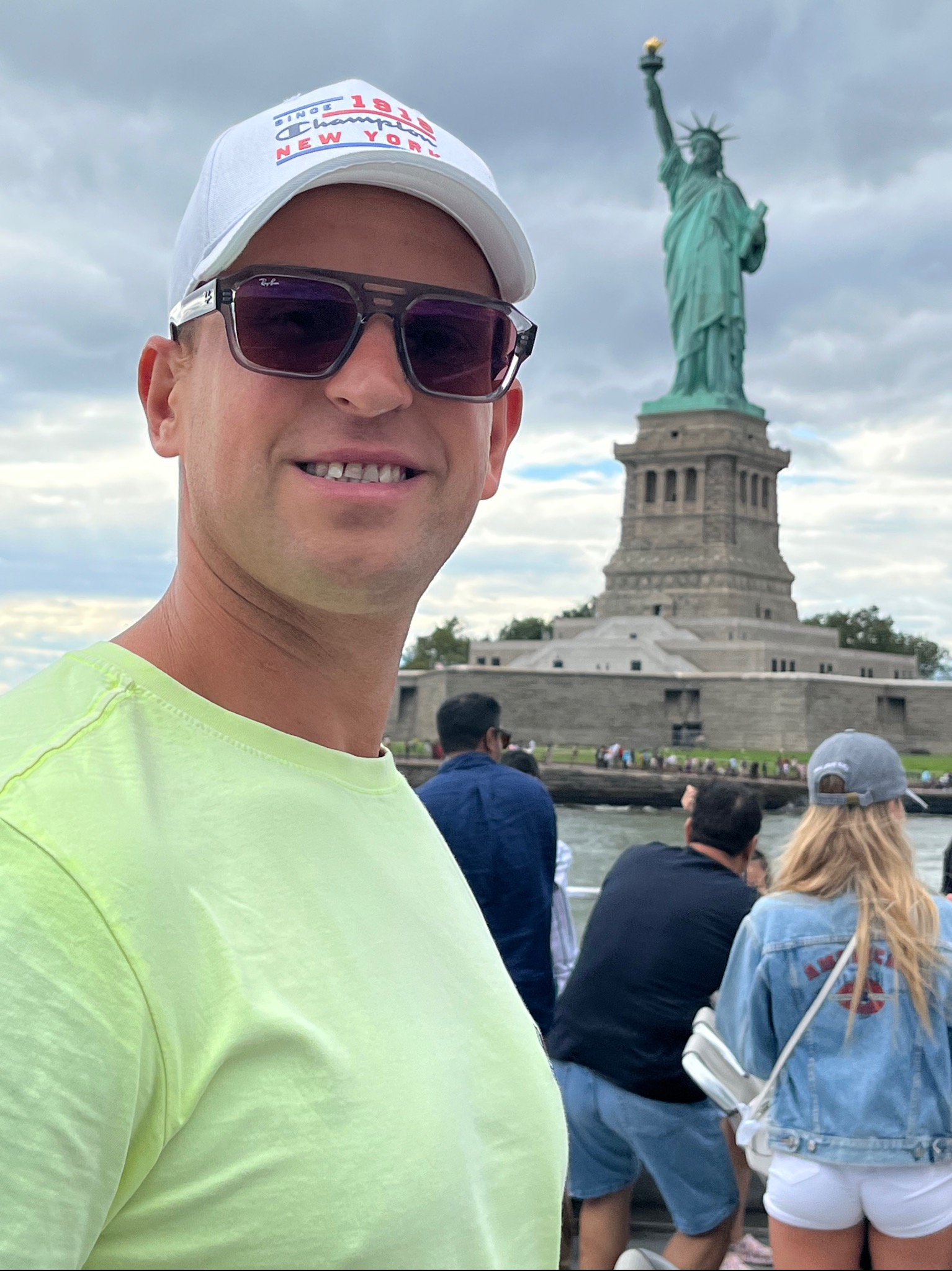 Uśmiechnięty mężczyzna w okularach i czapce na tle Statuy Wolności, widoczni inni turyści, pochmurne niebo.