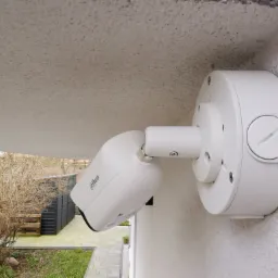 Biała kamera monitoringu Dahua zamontowana na elewacji budynku, widok z dołu, w tle ogród z krzewami i budynkiem gospodarczym.