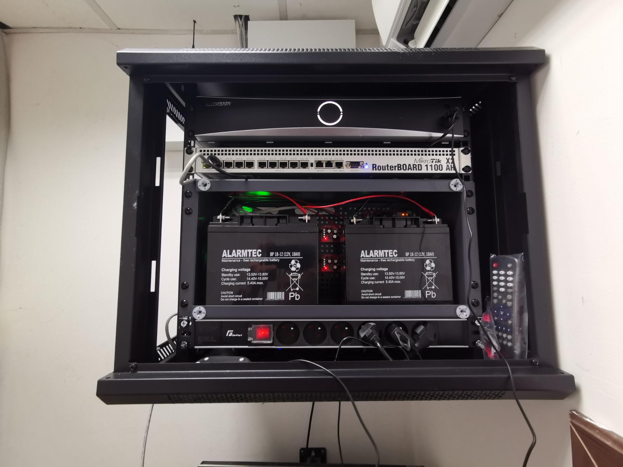 Szafa rackowa z urządzeniami: router MikroTik, zasilacz buforowy z dwoma akumulatorami Alarmtec, listwa zasilająca Gefort, kontroler Hikvision, przewody i kable.