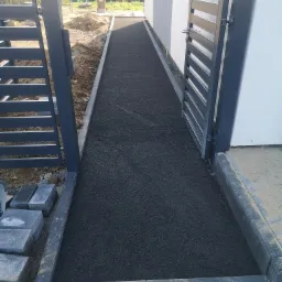 Ścieżka z drobnego, ciemnego kruszywa prowadząca od furtki do wejścia domu, obramowana szarą kostką i widoczne niedokończone prace ziemne po bokach.