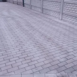Świeżo ułożona, jasnoszara kostka brukowa tworząca podjazd, obramowana kostką brzegową. W tle betonowe ogrodzenie z fakturą cegły.