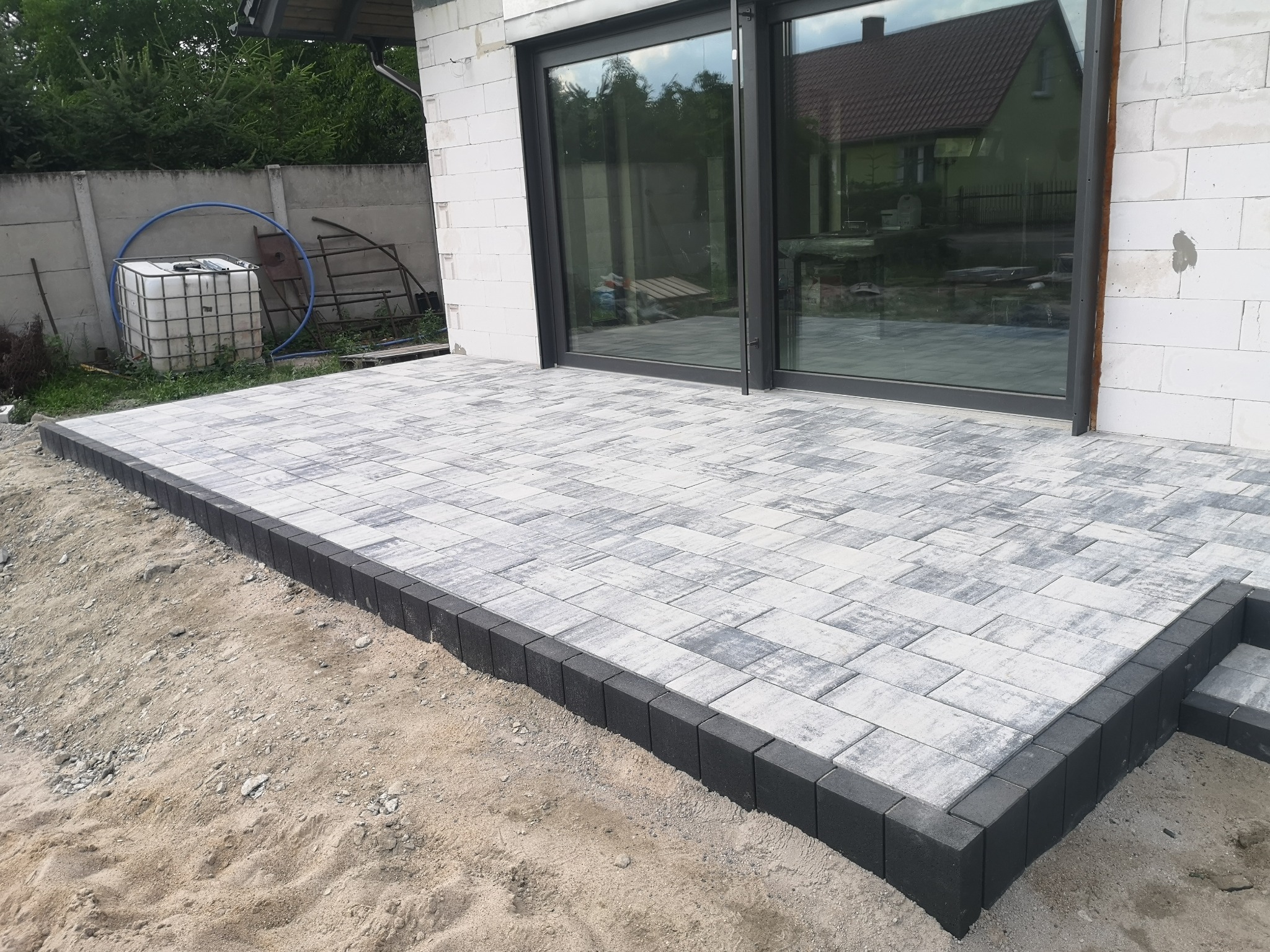 Nowo ułożony taras z jasnoszarej kostki brukowej z ciemnoszarym obrzeżem, widok z niskiej perspektywy, surowy stan terenu wokół, w tle biały budynek w stanie surowym i duże okno tarasowe.