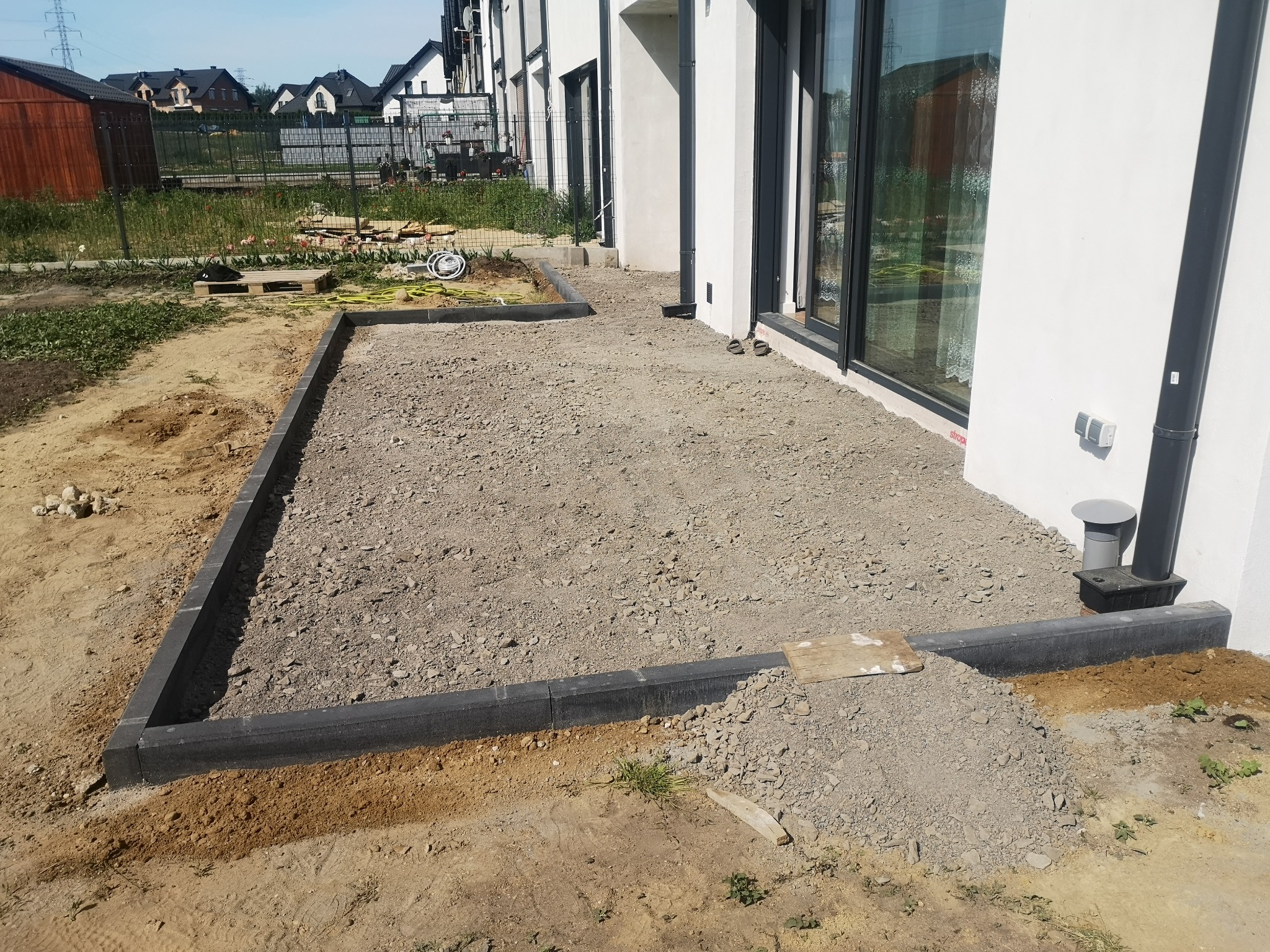 Przygotowany teren pod taras z obrzeżami z grafitowej kostki, podsypany żwirem, widok na nowoczesny dom z dużymi oknami.