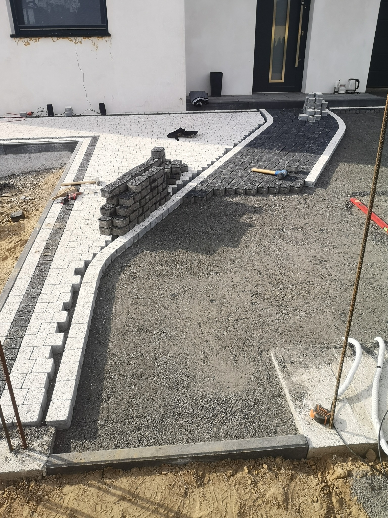 Fragment układanej ścieżki z kostki brukowej w dwóch kolorach, z widocznymi narzędziami i stosem nieułożonych kostek, na tle białego domu z czarnymi drzwiami.