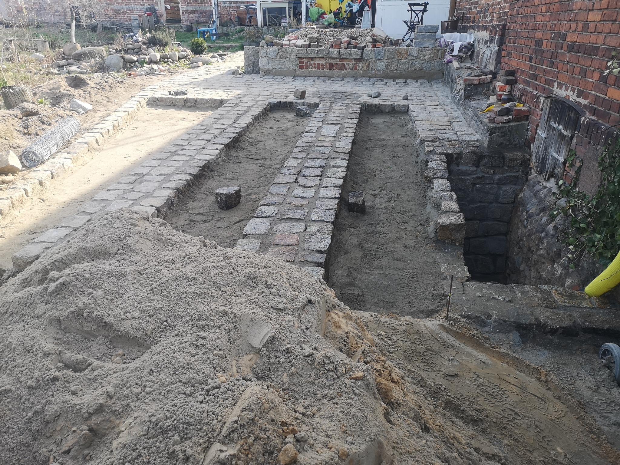 Przygotowanie podłoża pod ścieżkę z kostki granitowej, widoczne obrzeża z kostki, wykopane koryta i pryzma piasku.