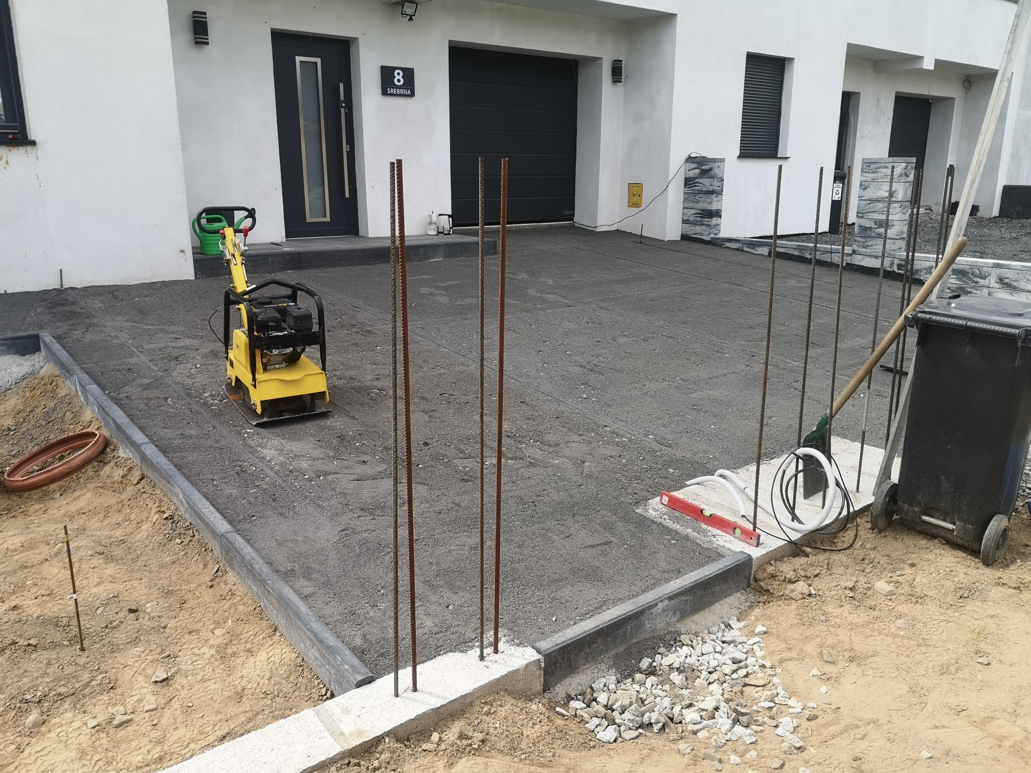 Żółta zagęszczarka obok domu z numerem 8 na podsypce piaskowej przed ułożeniem kostki brukowej, widoczne wystające pręty zbrojeniowe i obrzeża.